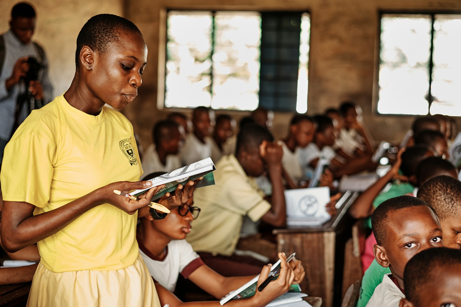 A teacher standing in a full classroom, checking a stack of exercise books by hand